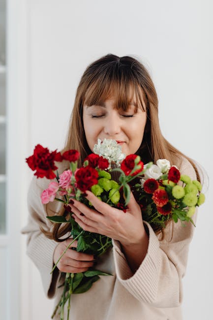 A woman with light brown, shoulder-length hair and bangs is holding a colorful bouquet of fresh flowers close to her face, with her eyes gently closed and a subtle smile. The bouquet includes vibrant 