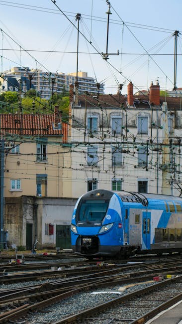 The image features a modern blue and gray double-decker train approaching a railway station, with overhead electrical lines and support structures visible above the tracks. In the background, there ar