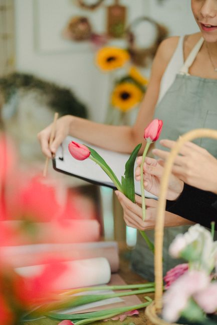 A person holding a floral arrangement featuring two pink tulips with green stems and leaves, inspecting or adjusting the bouquet with a smartphone in a brightly lit floral shop. The bouquet is partial