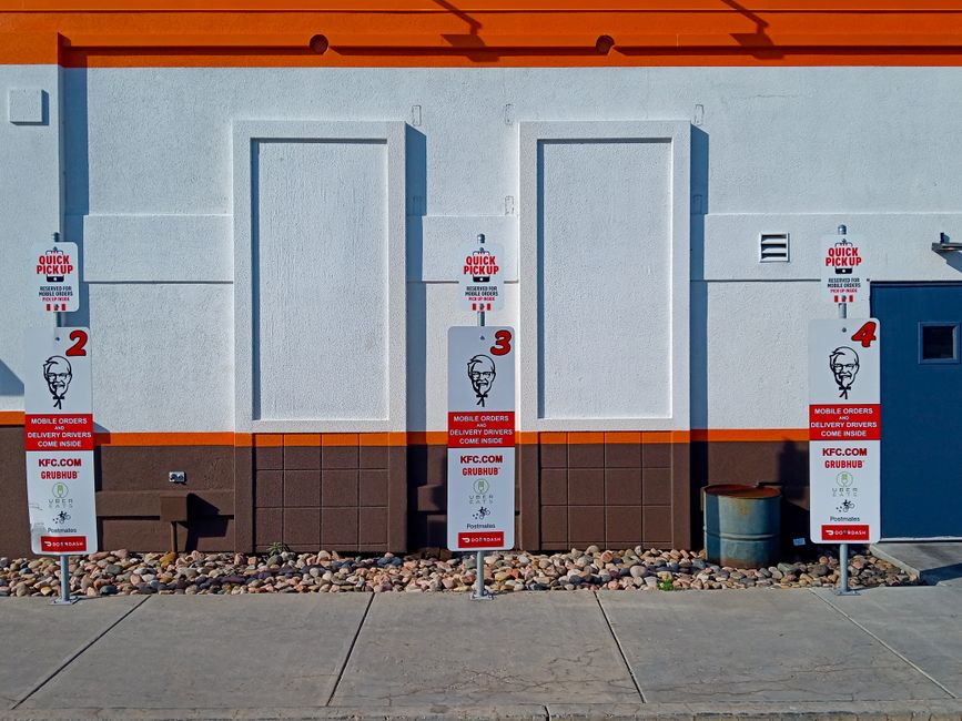 The image depicts the exterior wall of a KFC fast food restaurant, featuring three white, rectangular, blank panels on a light beige wall, each framed with orange trim at the top. In front of the wall
