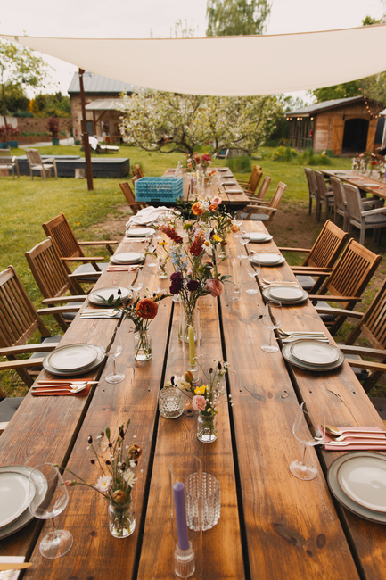 A long wooden outdoor dining table set for a special occasion, decorated with a variety of small glass vases filled with fresh, colorful flowers including daisies, roses, and wildflowers in shades of