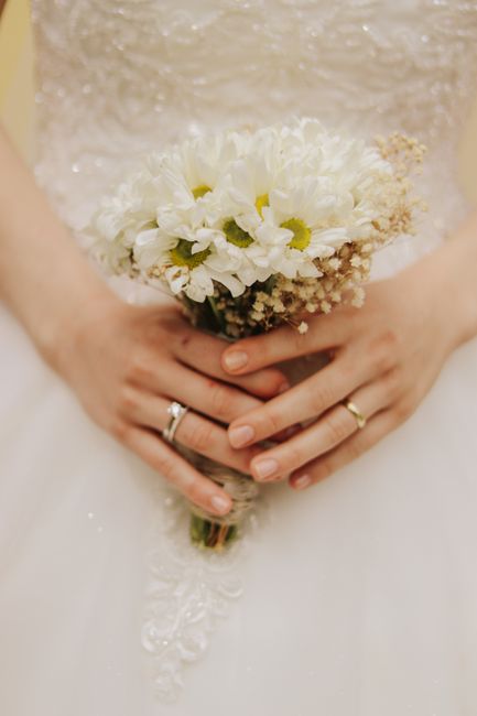 The image depicts a bride holding a bouquet of fresh flowers in front of her waist. The bouquet features white and cream-colored blooms, including large white roses or peonies, surrounded by smaller w