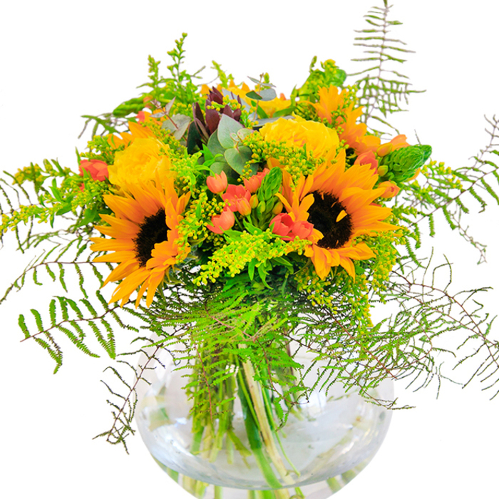 Radiating exuberance with every bloom, this handcrafted bouquet combines the classic charm of the Harpenden area with a riot of colour and texture. At the heart of the arrangement, large, radiant sunflowers dazzle with their golden petals and pronounced brown centers-evoking happy memories of summer fields by the River Lea. Interlaced among the sunflowers, buttery yellow roses spiral open, their intricate layers lending a softness and sophistication reminiscent of old gardens along Station Road. Scattered boldly within the bouquet, shiny red Hypericum berries pop against a rich tapestry of greenery: cascades of soft fern fronds, spiky green fillers, and accent sprays of tiny yellow goldenrods mirror the untamed elegance of Aldwickbury parks. Sprigs of blue-green eucalyptus impart a cool note, balancing the warm hues and giving the design a fresh, rustic finish. Nestled within a clear round vase-its transparent walls revealing the intertwining stems and glimmering water-the flowers come alive in the gentle light, their textures and colours inviting touch and admiration. The subtle fragrance of fresh-cut blooms drifts in the air, stirring feelings of joy and nostalgia, as though bringing a little of Harpenden's countryside into any home. Perfect for festive occasions or spontaneous gifting, this arrangement stands as a tribute to vibrant moments and local florist artistry.